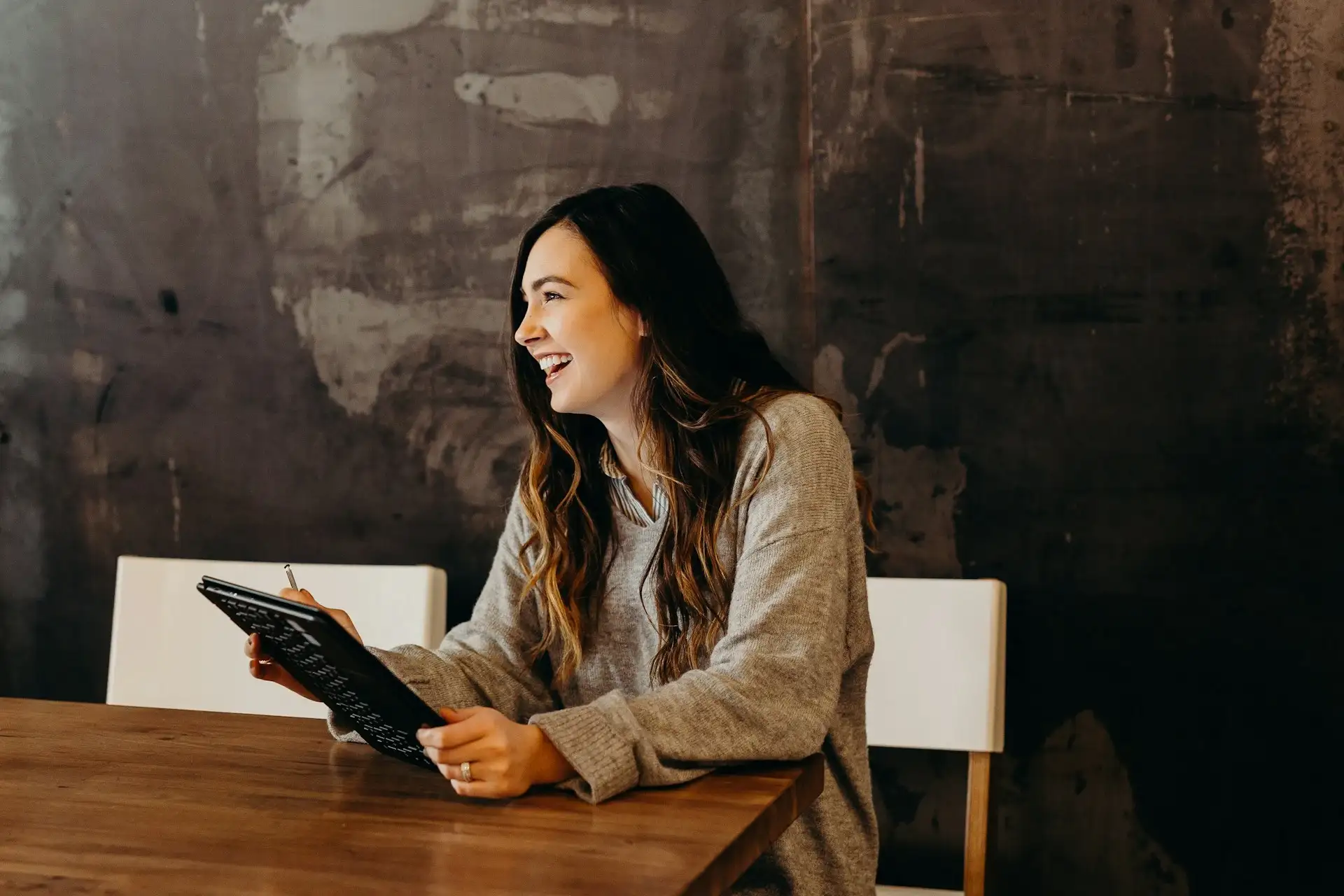 A smiling girl sits at a desk, holding a tablet in her left hand and a stylus in her right hand. She looks away from the tablet, seemingly amused by something out of view. Her expression suggests enjoyment and engagement with the content.