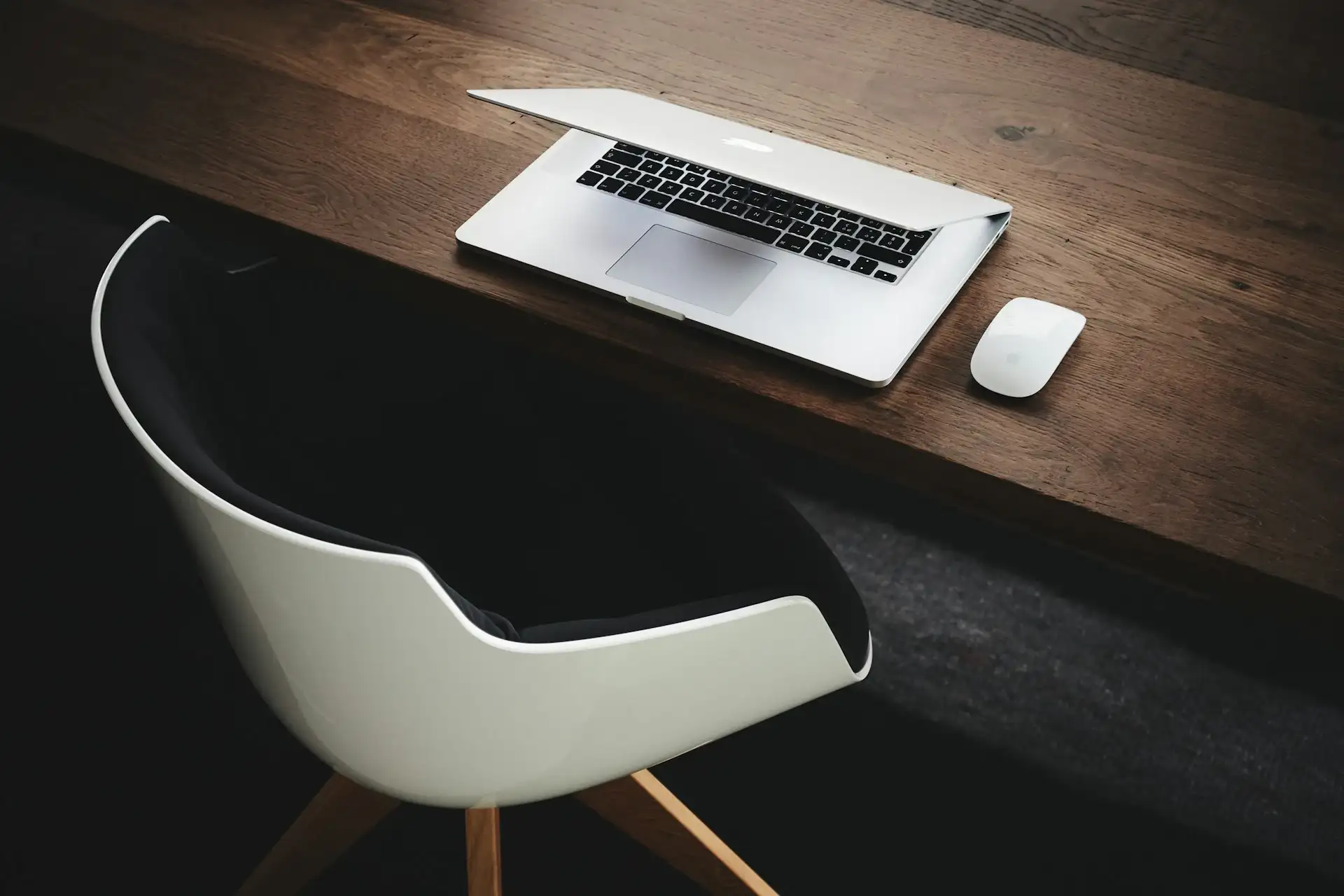 A clean and tidy desk with a chair. An Apple laptop rests on the desk, half-opened, next to a wireless mouse. The setup suggests an organized workspace ready for use.
