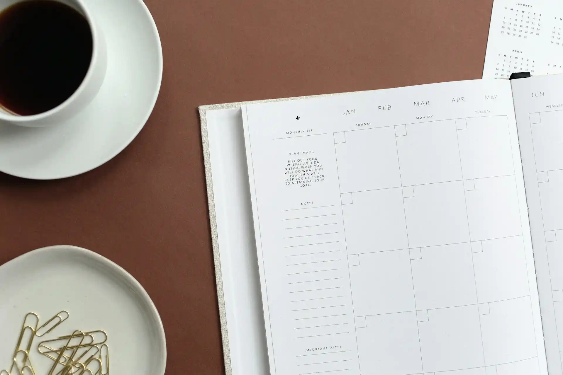 Top view of a table with an open blank monthly calendar as the focal point. A cup of coffee is placed at the top of the calendar, and some paper clips are to the left.