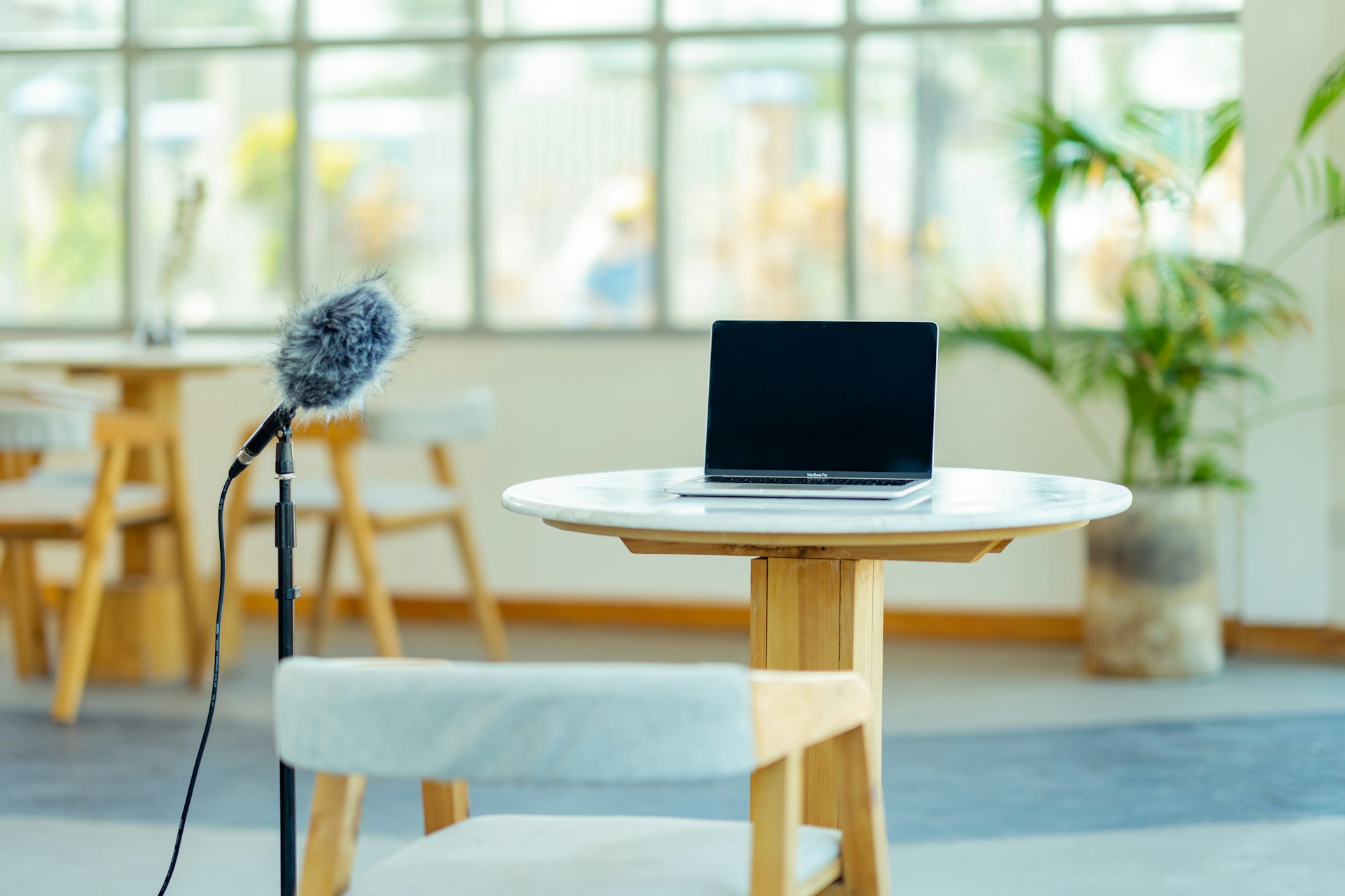 A photo of a common area with a table and three chairs in the background, along with a plant to the right. In focus is a table with a single chair, featuring an open laptop and a microphone positioned on the left side.