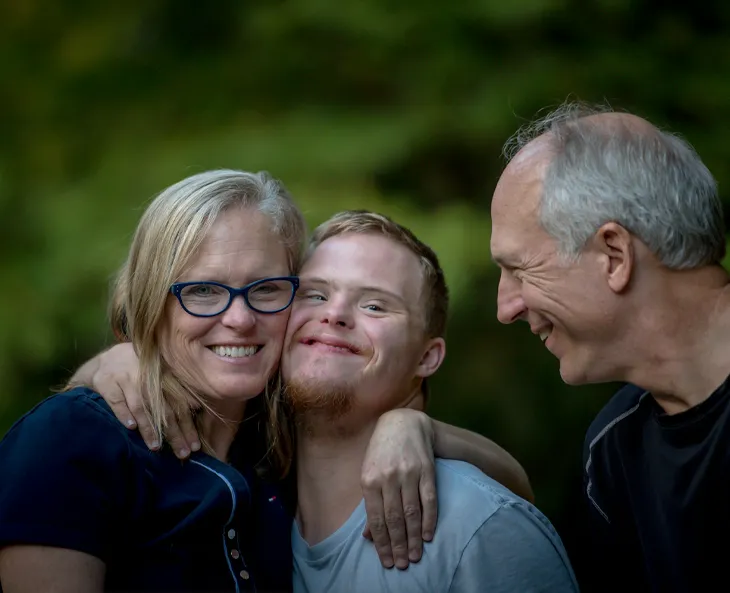 A smiling family portrait where the mom has her arm around her disabled son, who is grinning widely. The son reciprocates by placing his arm around his mom. The dad, also smiling, gazes affectionately at both his wife and son."