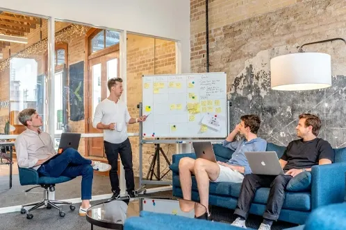 Group of 4 people creating a plan, three of them are sitting down and one is standing up pointing to sticky notes on a dry erase board.