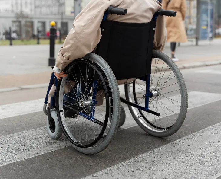 A close up image of a wheelchair with a person sitting down in the wheelchair, who is crossing the road at crosswalk intersection.