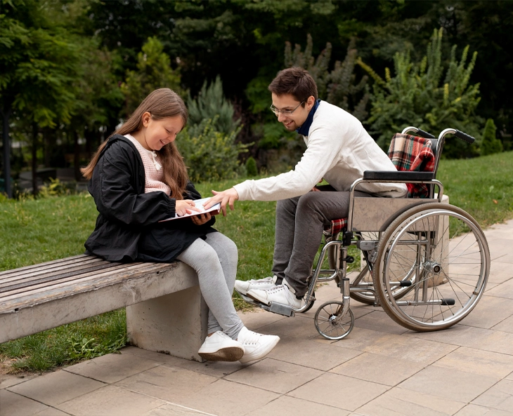 An image of a disabled person in a wheelchair, reading a book. The book is being held by a teenager who is also reading the book while sitting on a bench.